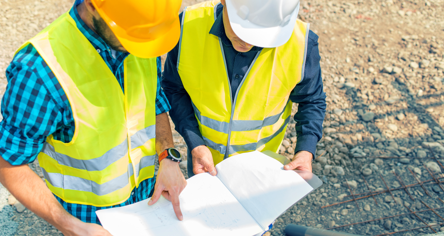 Two construction workers in safety vests and helmets reviewing blueprints at an active job site